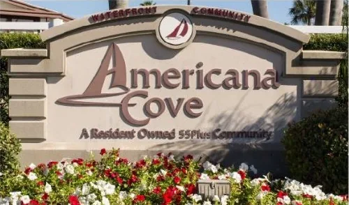 The entrance sign for Americana Cove, a resident-owned 55-plus community, framed by red and white flowers